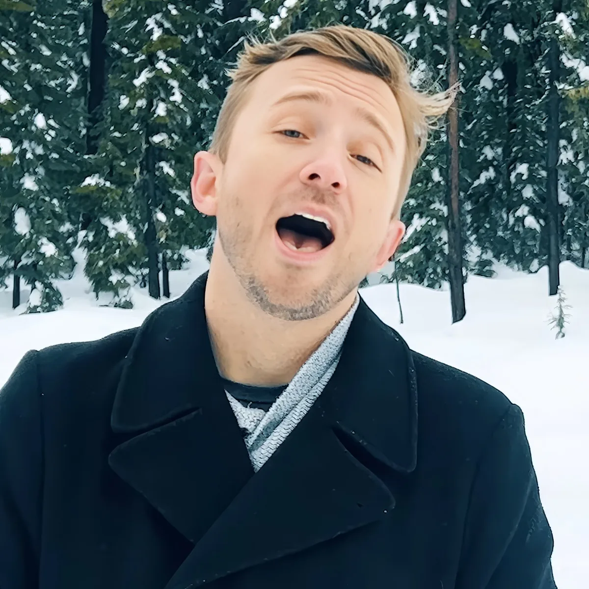 A man in a black coat and scarf stands in a snowy forest, mouth open, with snow-covered trees behind him.