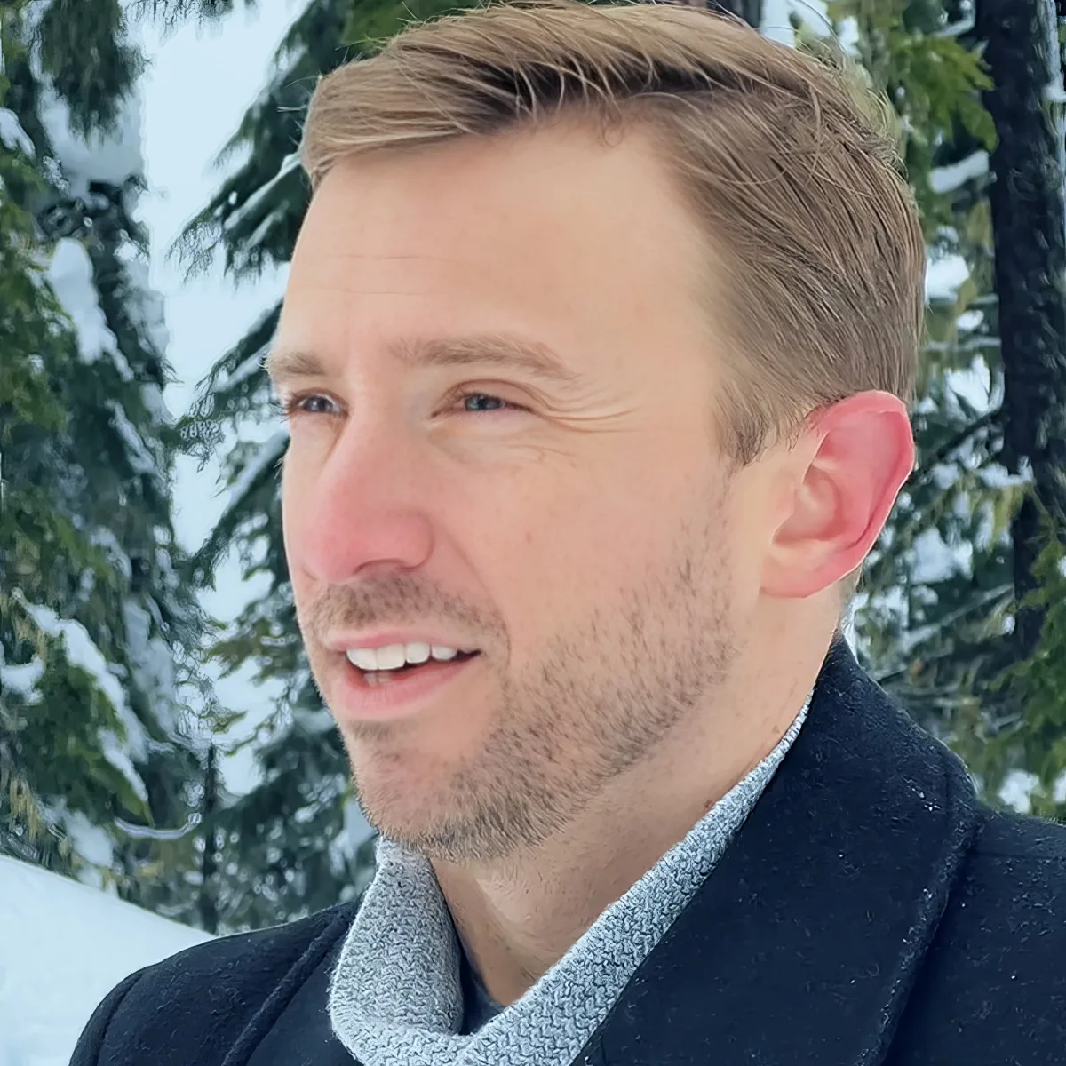 A man with short brown hair, wearing a dark coat and a light scarf, smiles in front of a snowy forest backdrop.