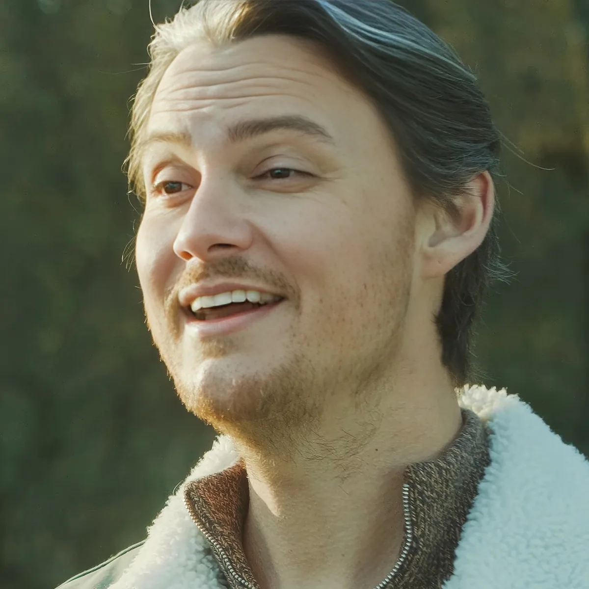 A smiling man with long hair and a beard, wearing a white and brown jacket, stands outdoors with a blurred green background.