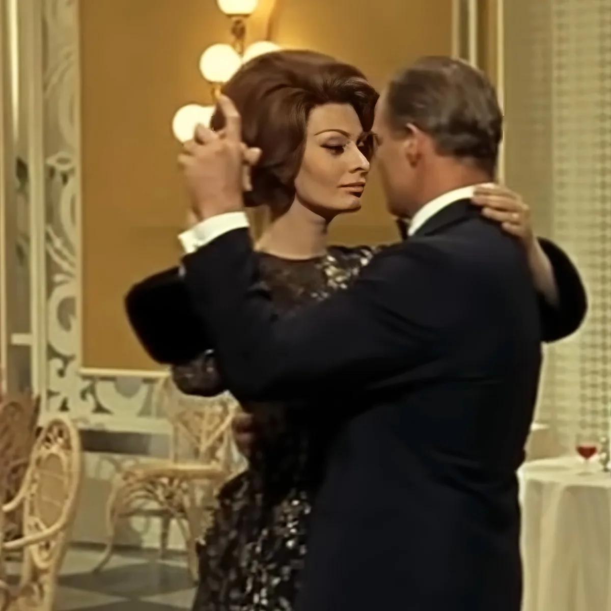 A man in a suit dances with a woman in a black sequined dress in a warmly lit room.