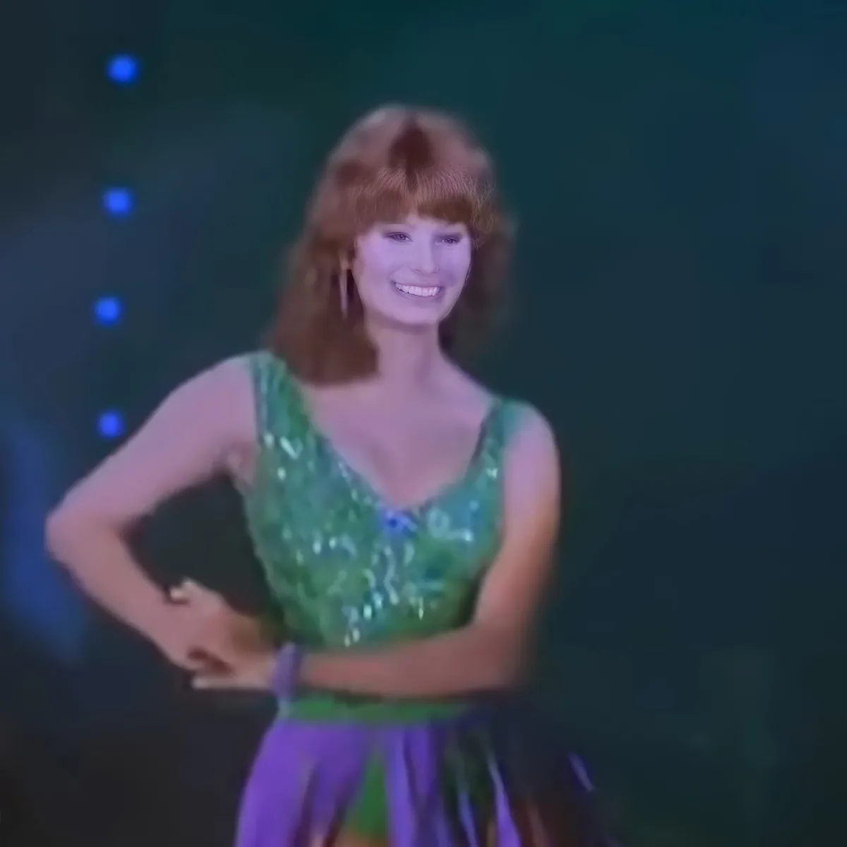 A woman in a green sequined top and purple skirt smiles with one hand on her hip.
