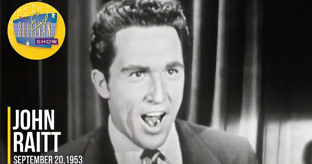 A man in a suit and tie is smiling widely with arms outstretched, possibly singing or speaking.