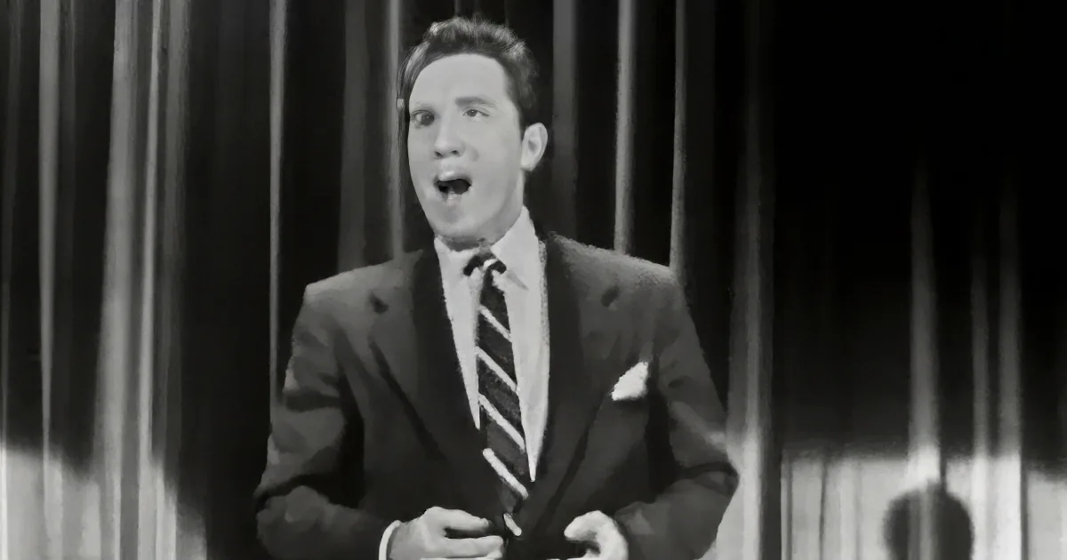A man in a suit and tie stands against a curtain backdrop, mouth open as if singing or speaking.