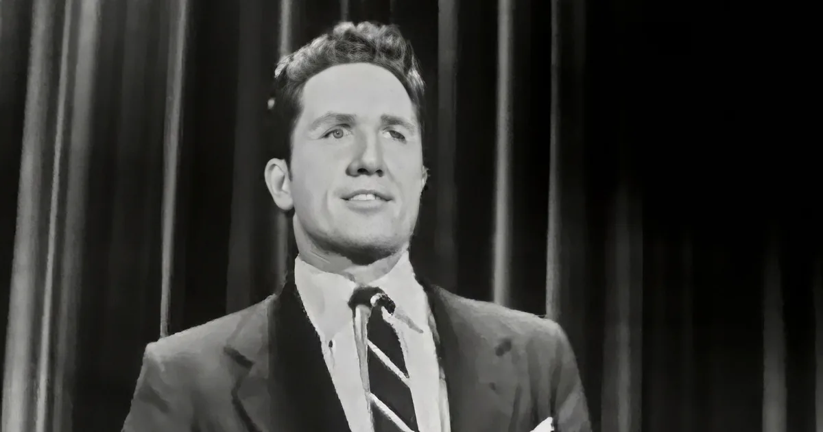 A man in a suit and tie stands in front of a curtain, looking up.