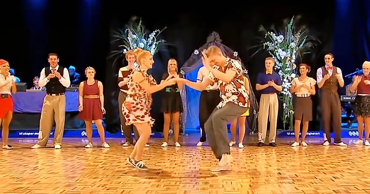 A couple dances on stage, surrounded by clapping spectators and floral decorations.