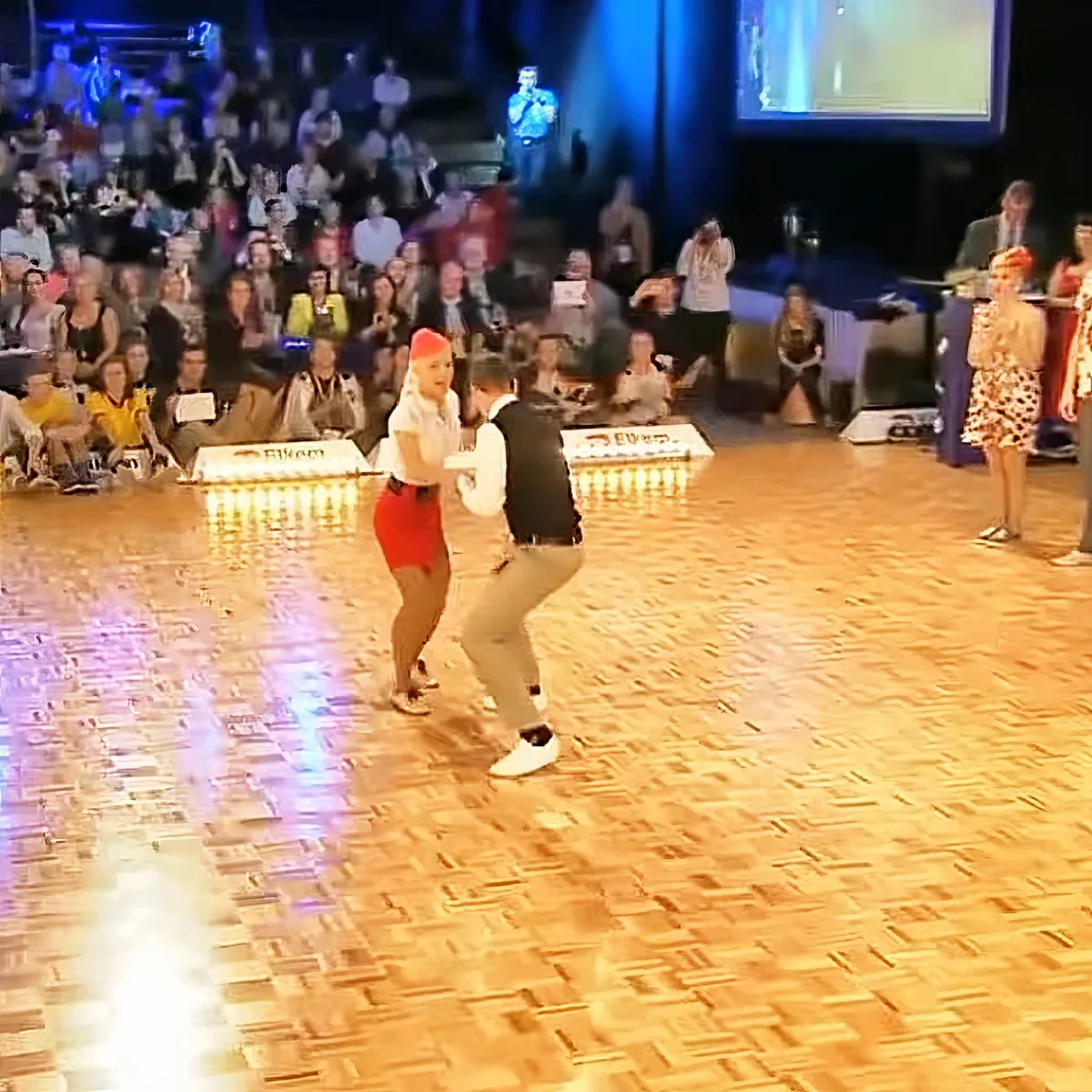 A couple dances on a wooden floor in front of an audience, with a person on stage and a screen in the background.