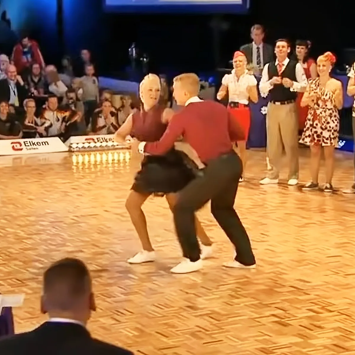 A man in a red shirt and black pants dances with a woman in a red top and black skirt on a wooden floor, surrounded by...