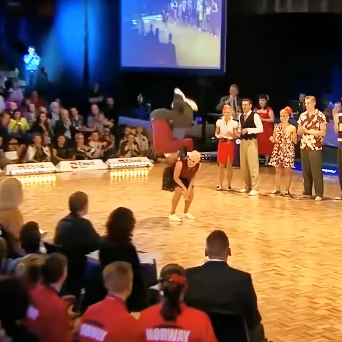 A person in a red outfit bends over on a stage, surrounded by spectators and performers.