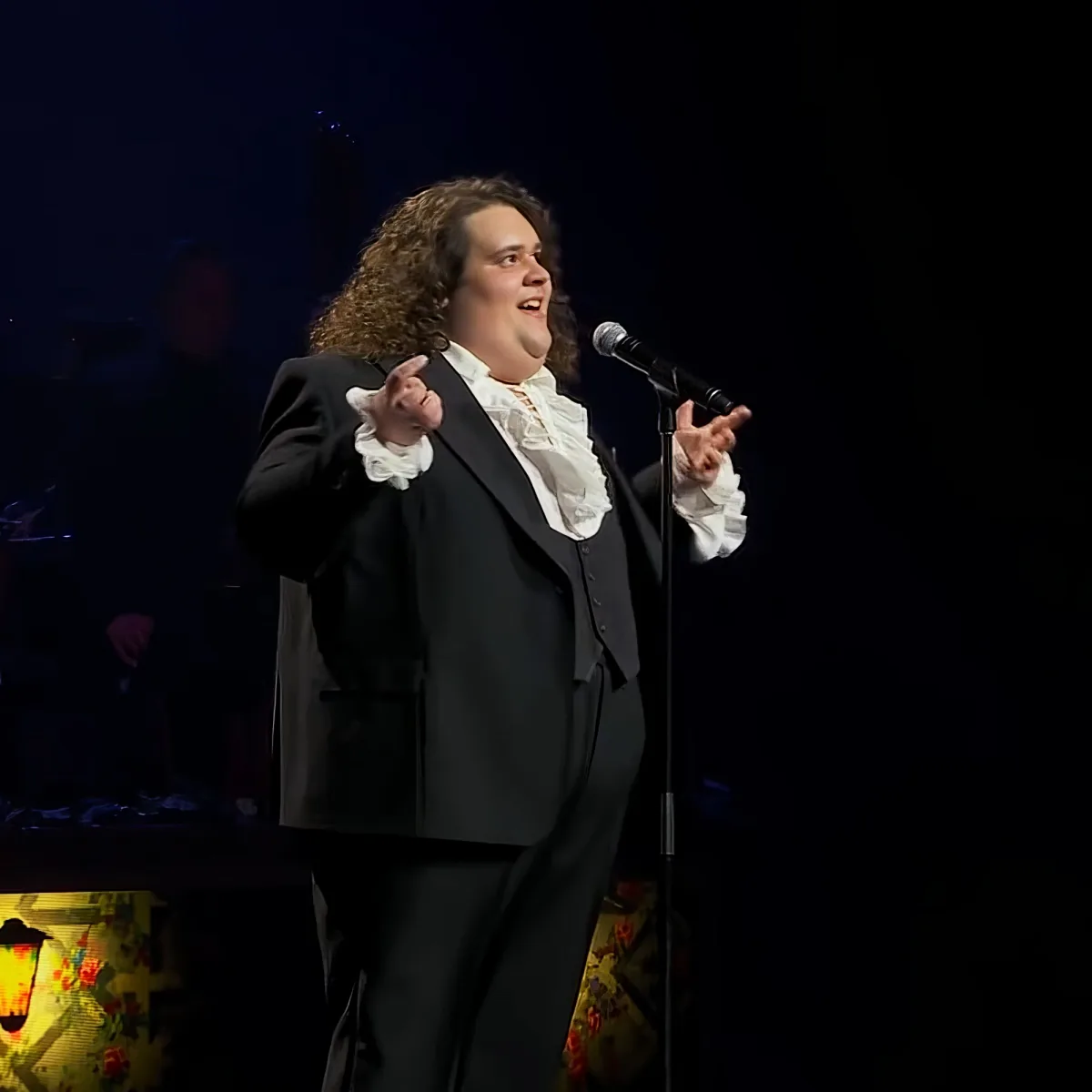 A person in a black suit and white ruffled shirt sings into a microphone on stage.