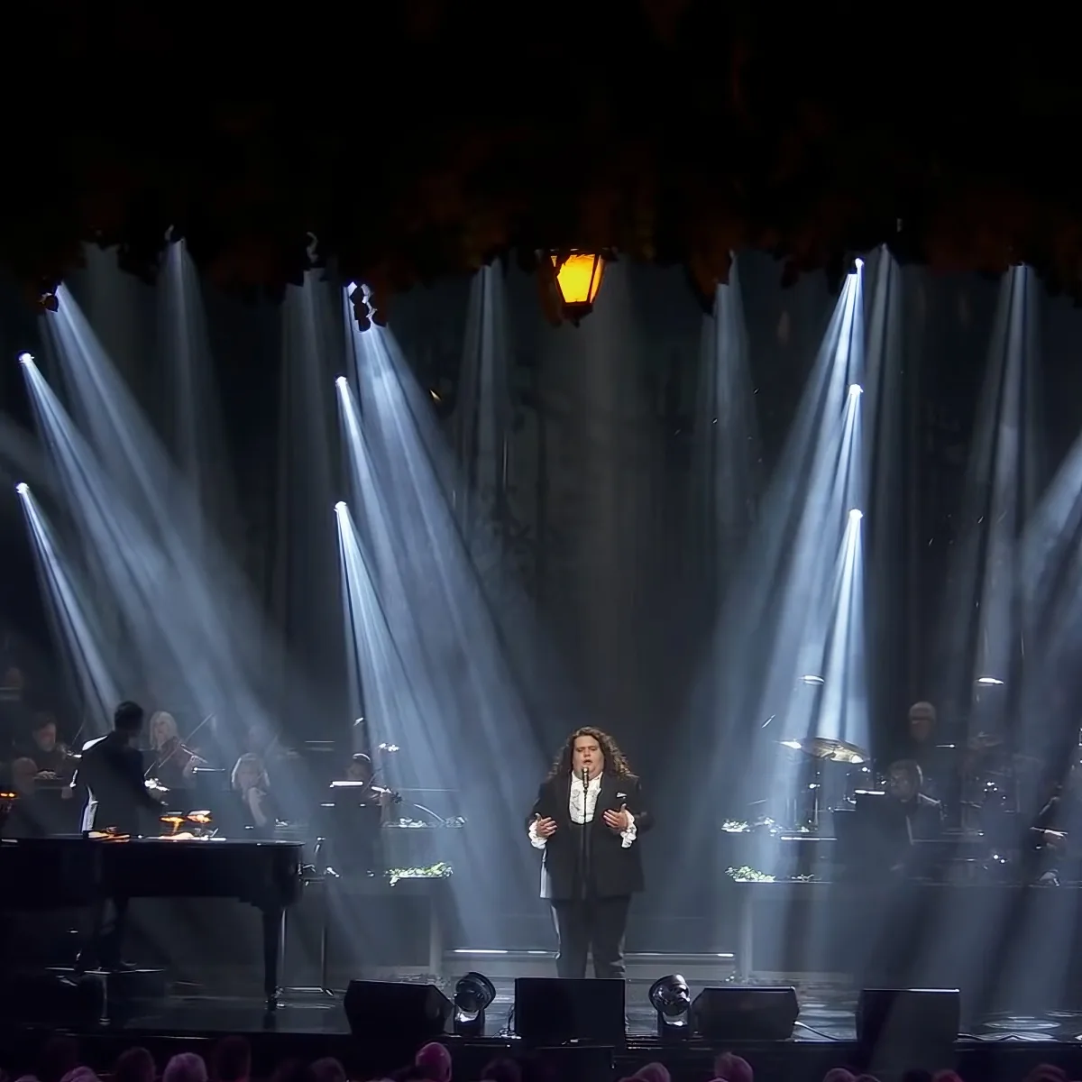 A person sings on stage with a band, illuminated by dramatic spotlights and a hanging lantern.