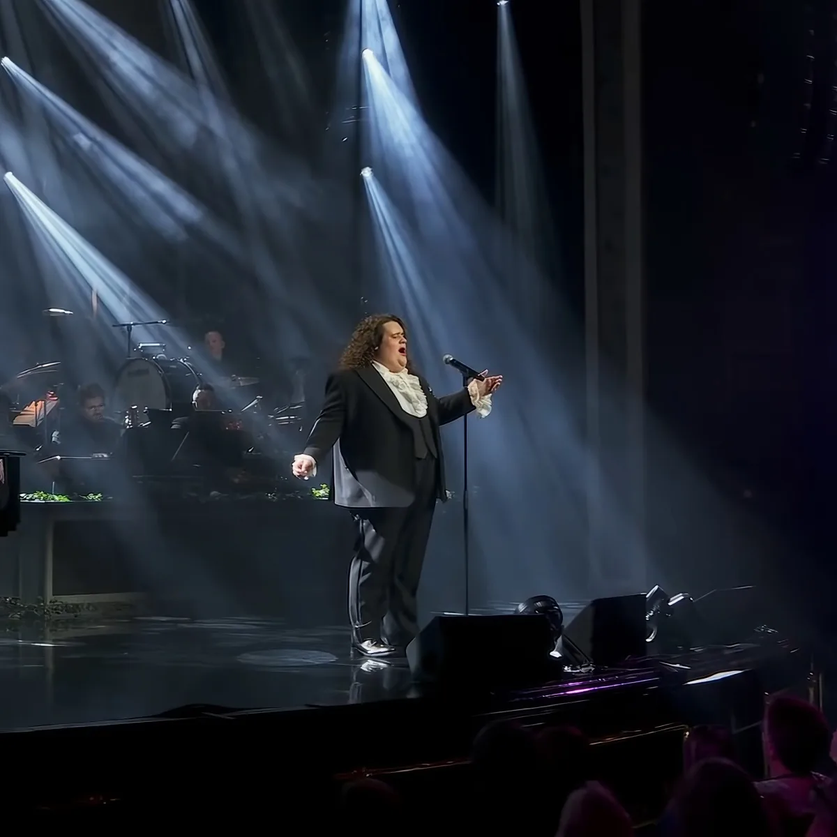 A person in a suit sings on stage with dramatic lighting and a band in the background.