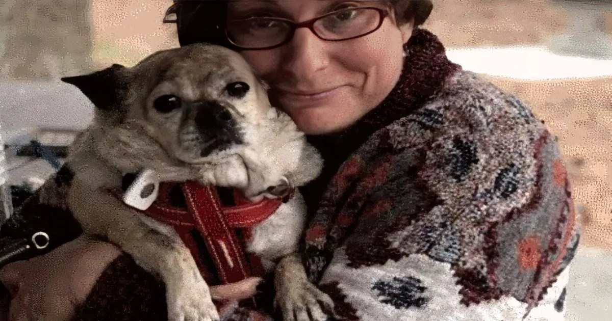 A woman kisses a small dog wearing a red harness in a park setting.