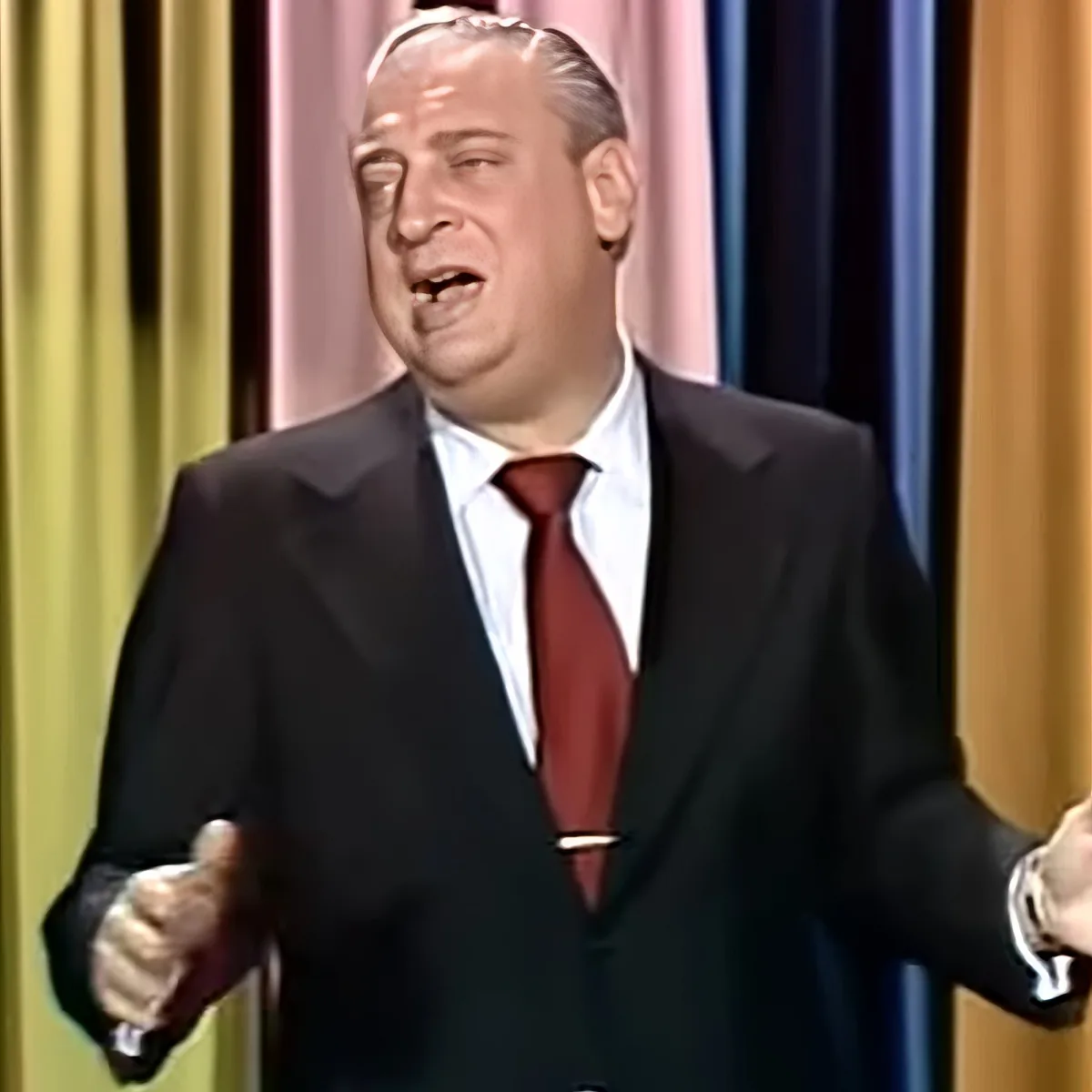 A man in a black suit and red tie stands with arms outstretched, possibly speaking or performing.