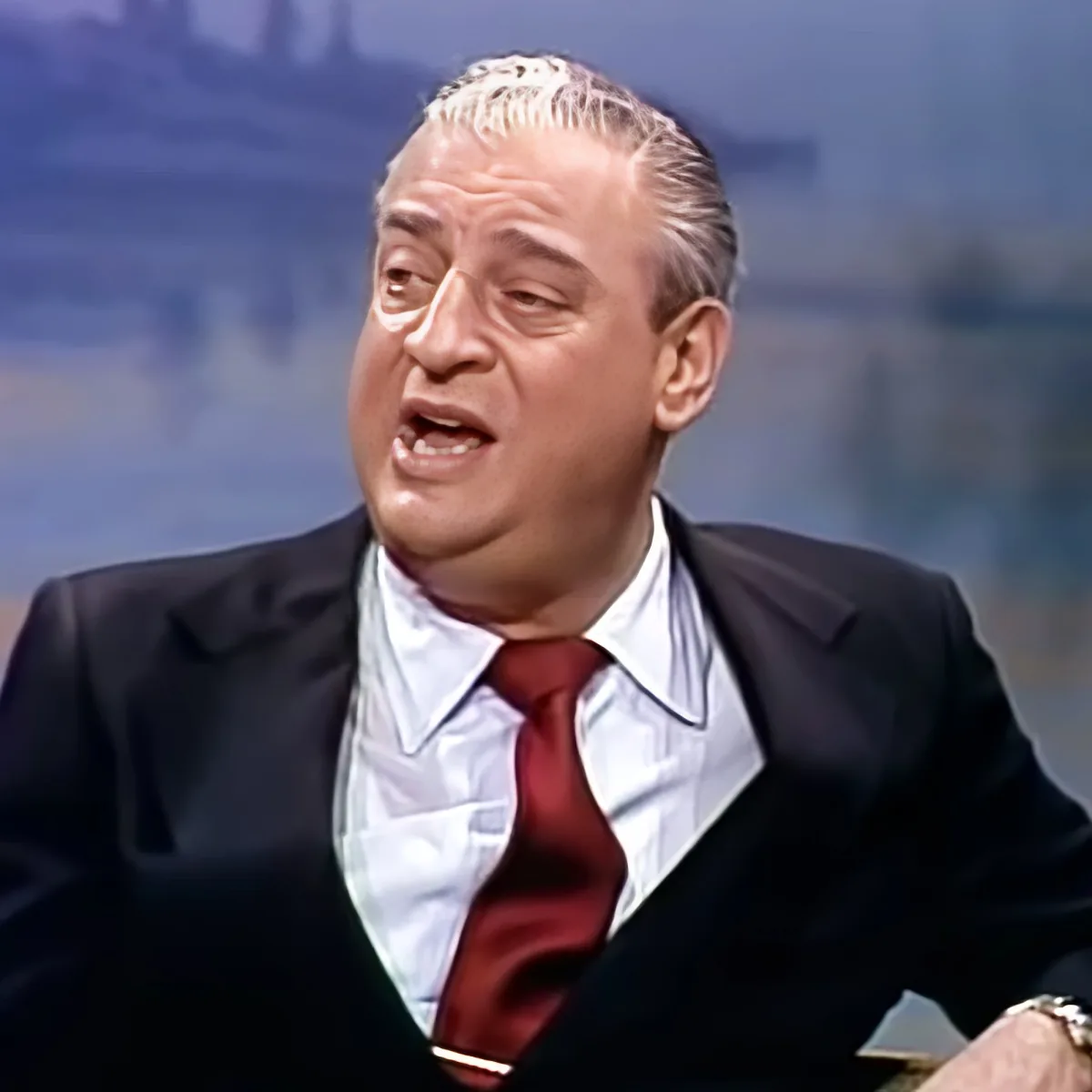 A man in a suit and red tie is speaking, with a cityscape in the background.