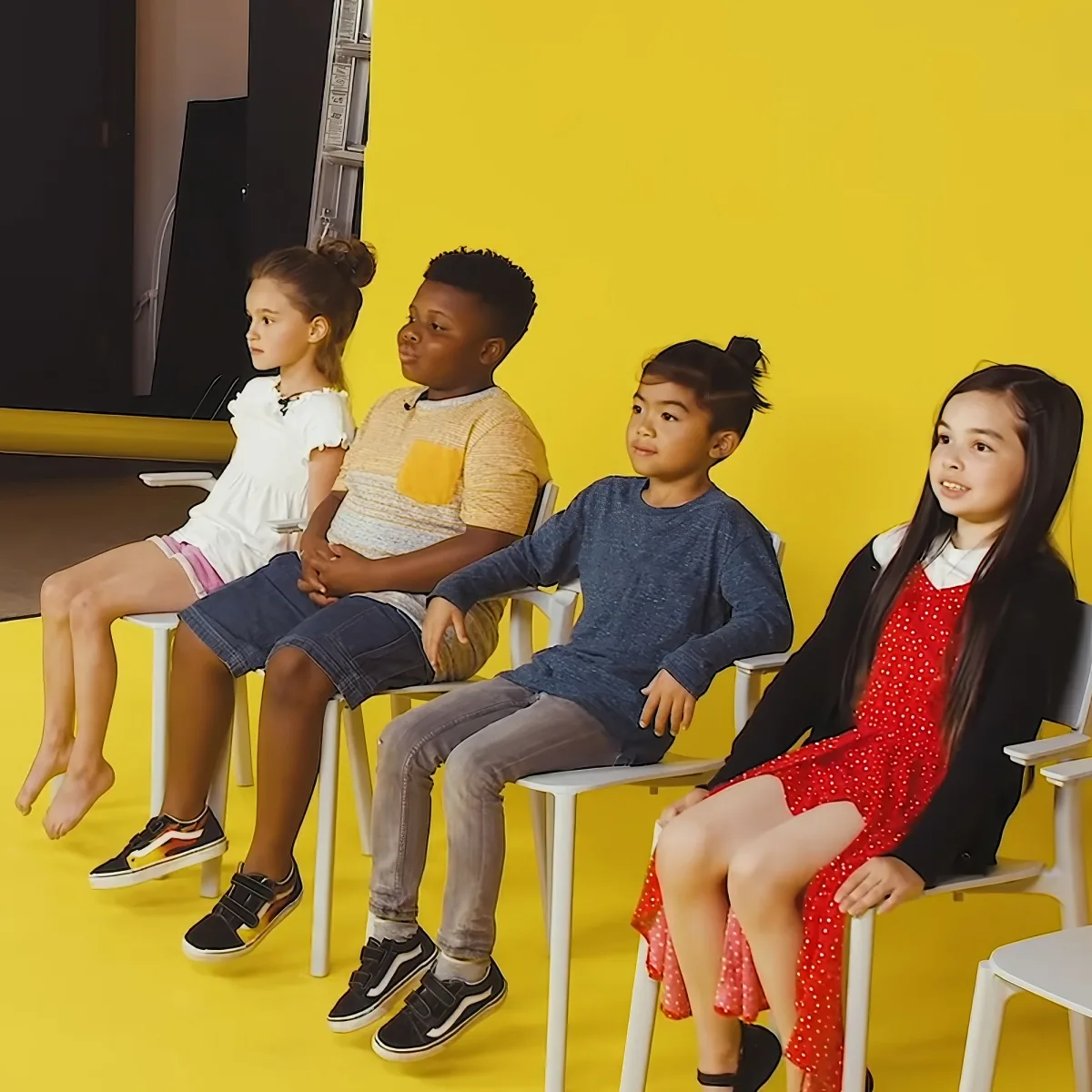 Four children sit on white chairs against a bright yellow wall, dressed in colorful outfits.