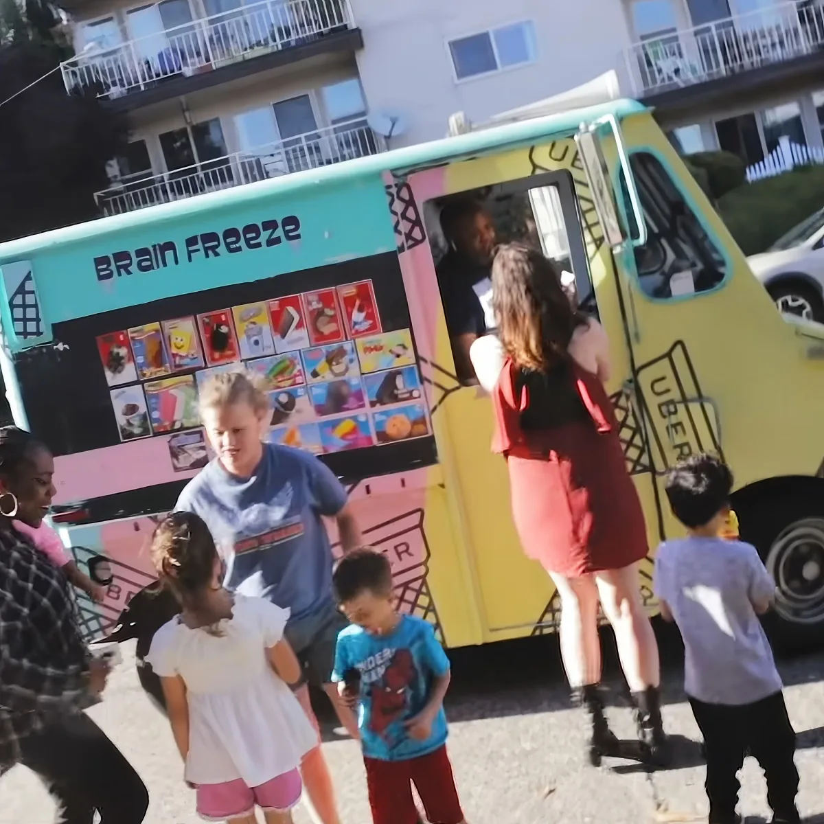 A woman in a red dress stands at a Brain Freeze ice cream truck, surrounded by children and a man in a blue shirt.