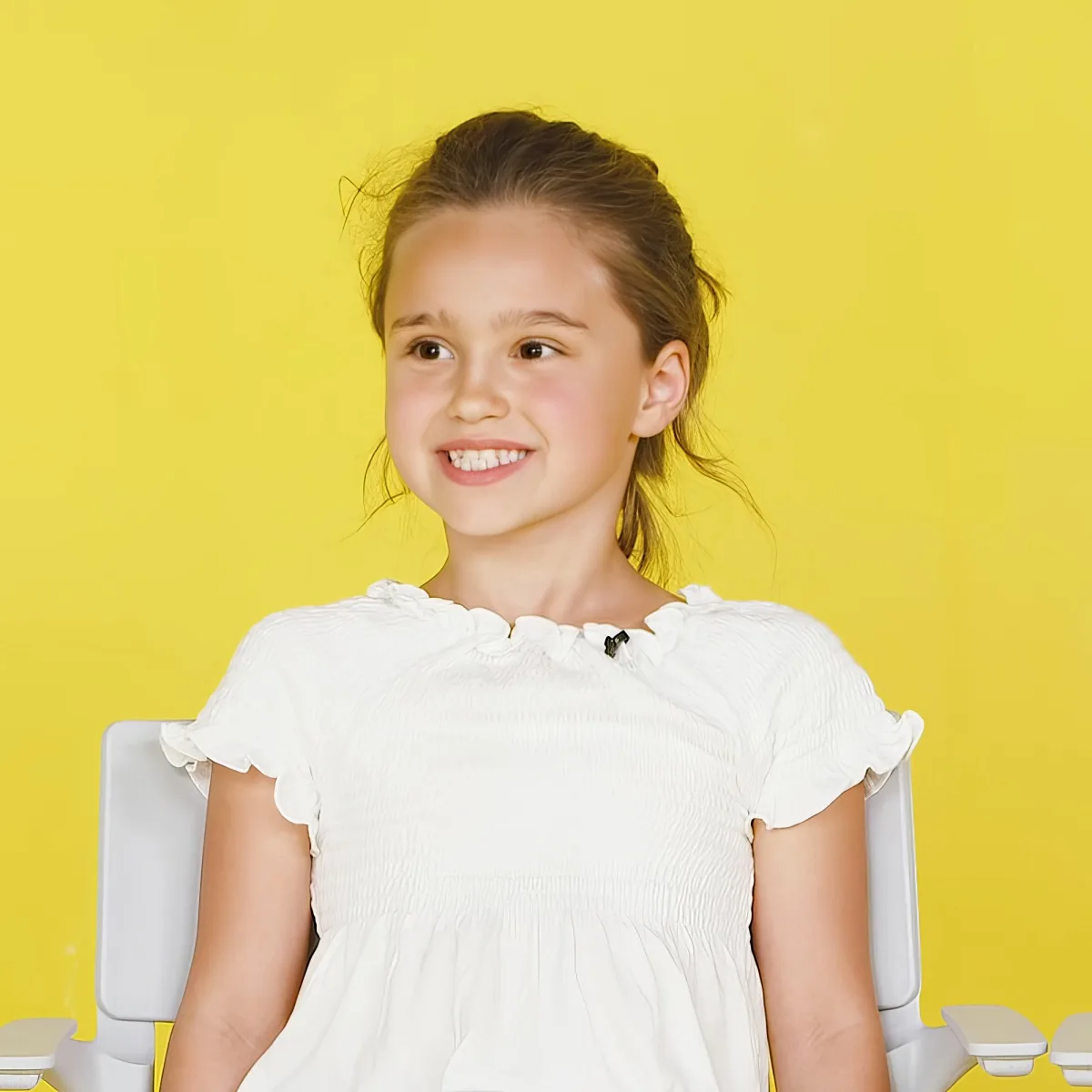 A young girl in a white dress sits against a bright yellow background, smiling.
