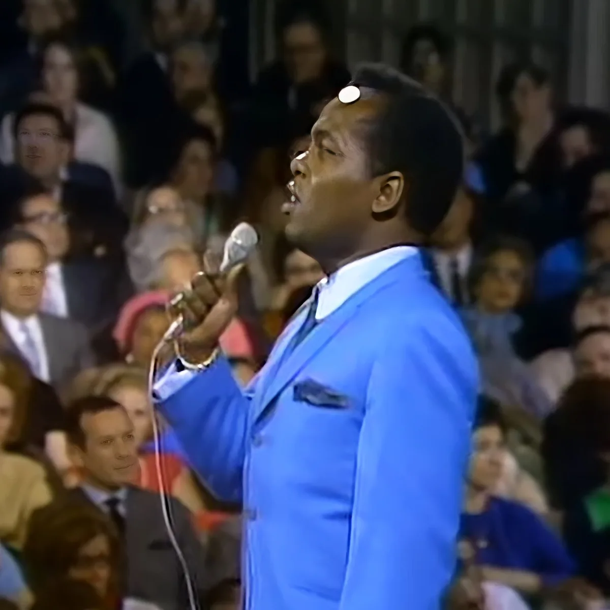 A man in a blue suit sings into a microphone in front of a crowd.