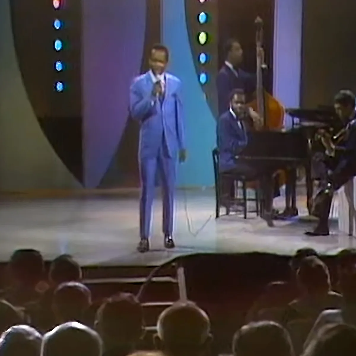 A man in a blue suit sings on stage, accompanied by musicians and a colorful backdrop.