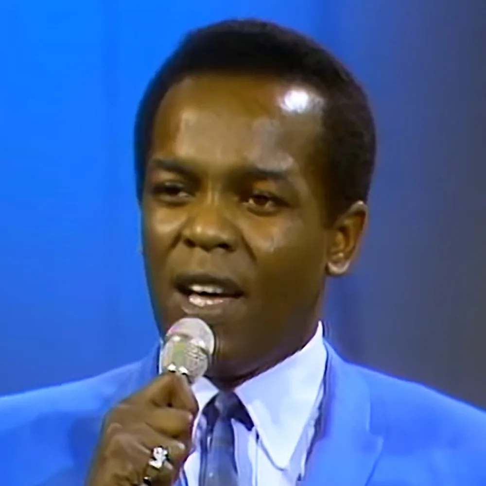 A man in a blue suit and tie holds a microphone, speaking with a blue background.