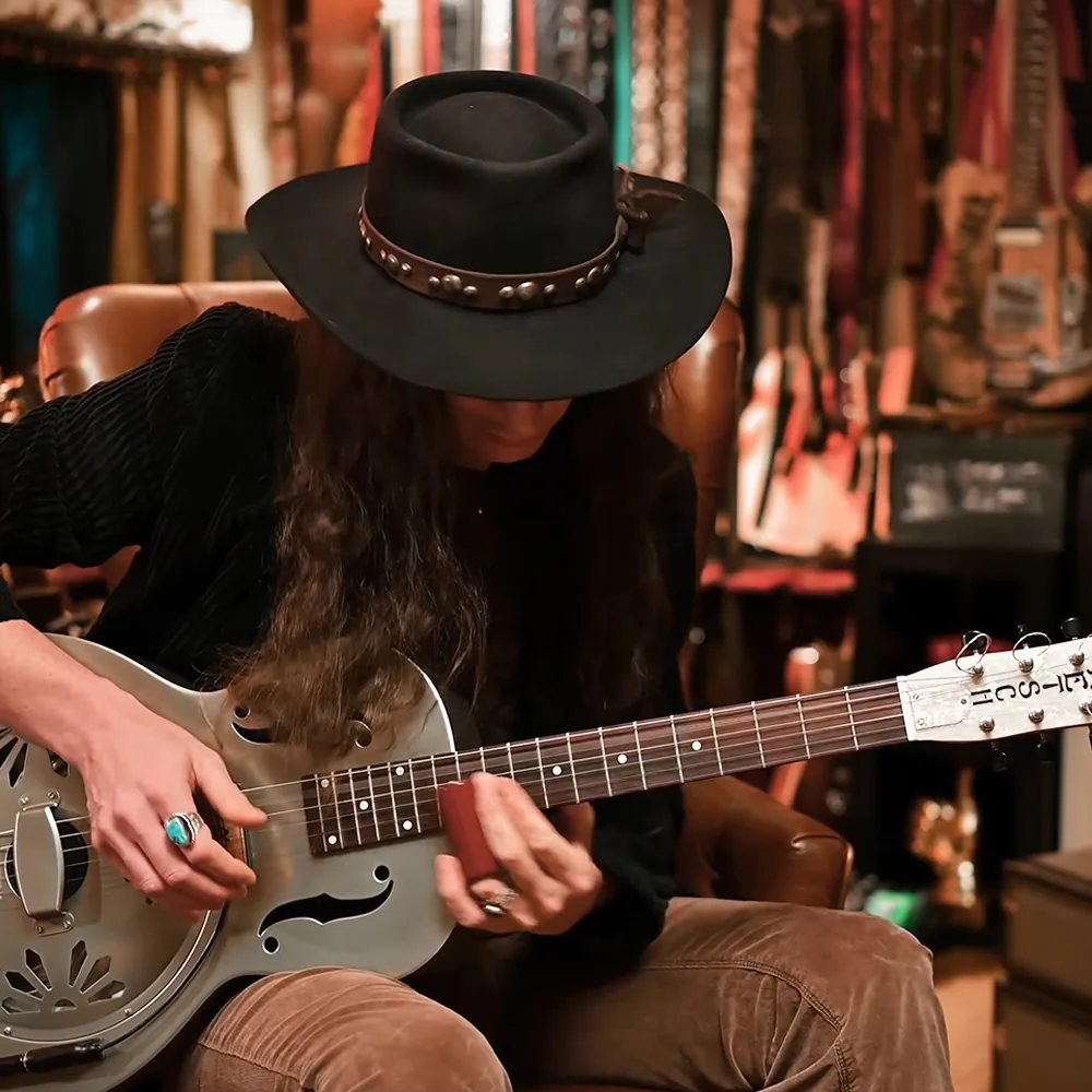 A person in a black hat and sweater plays a resonator guitar in a guitar shop.