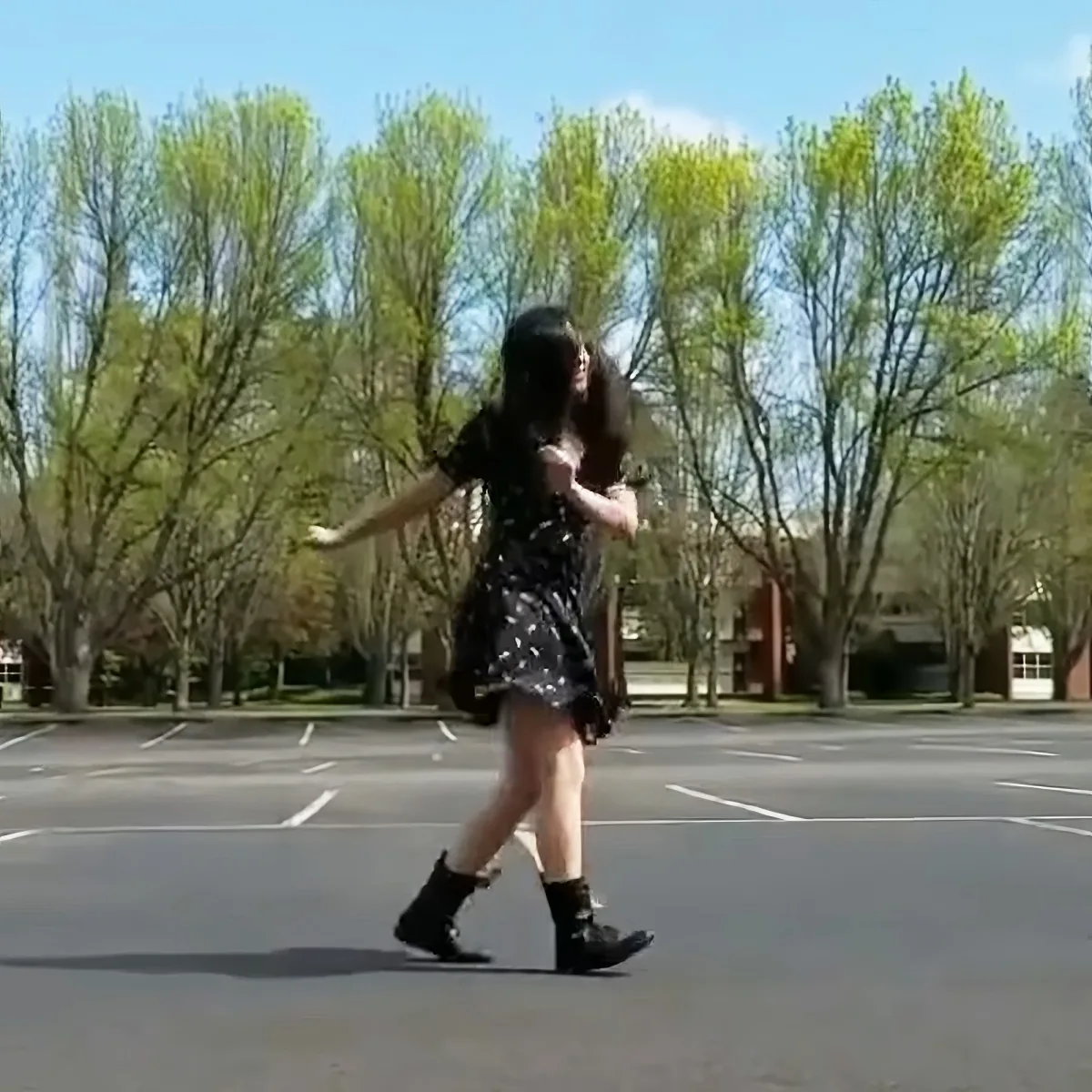 A person in a black dress and boots walks across a parking lot with trees in the background.