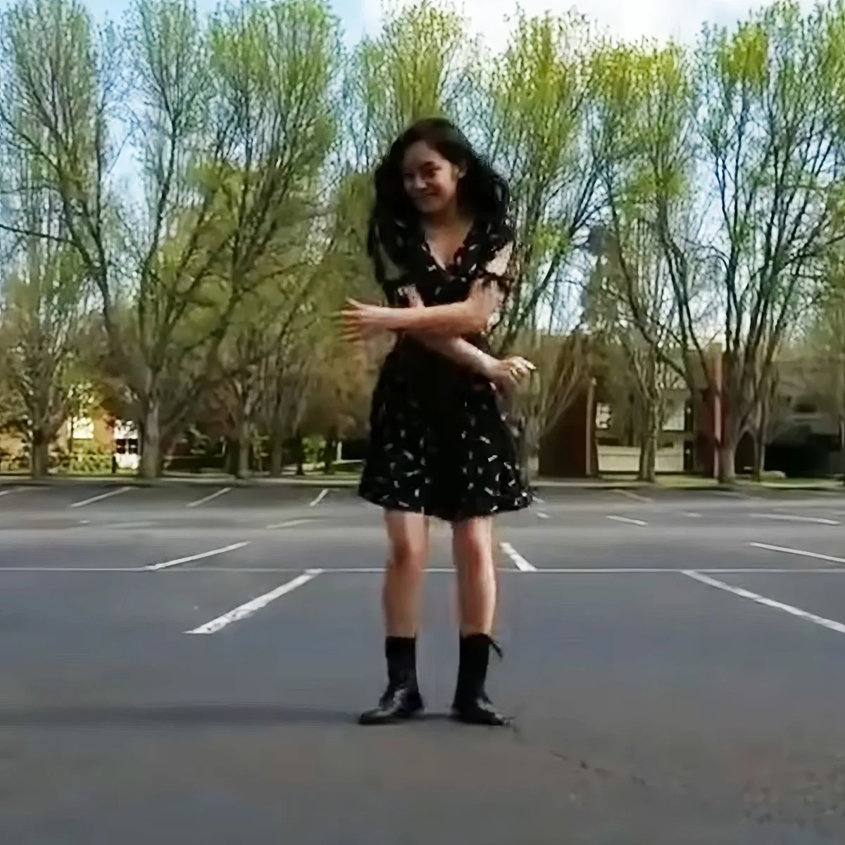 A person in a black dress and boots stands in a parking lot with trees and buildings in the background.