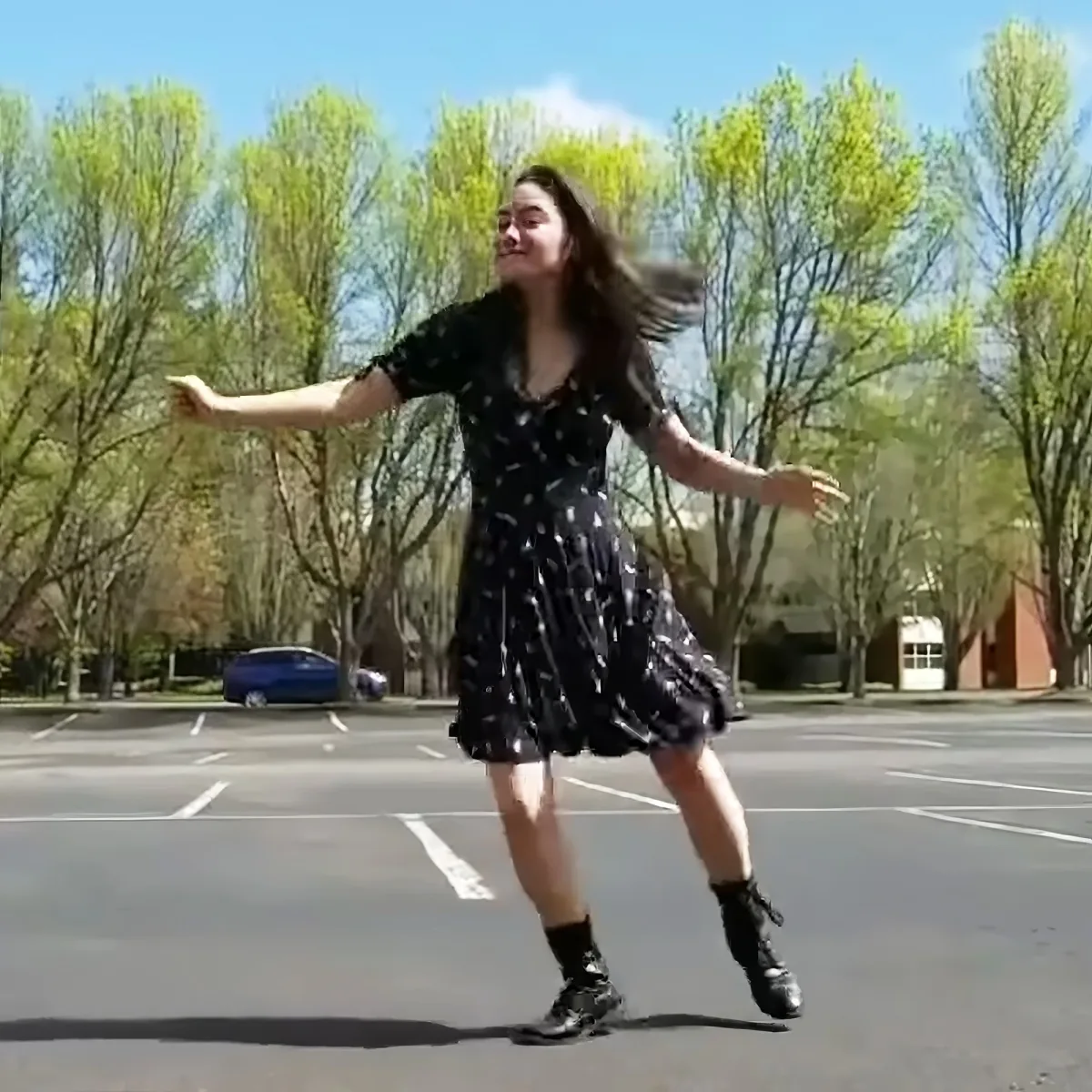 A person in a polka-dot dress and boots dances joyfully in a sunny parking lot with trees in the background.
