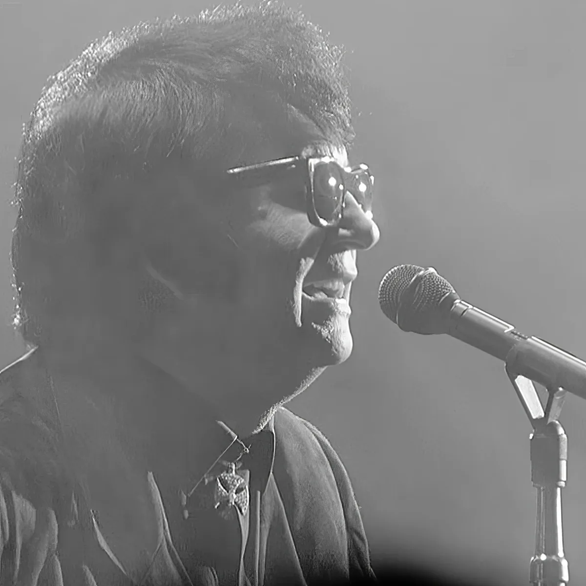 A person wearing glasses and a shirt with a bow tie sings into a microphone.