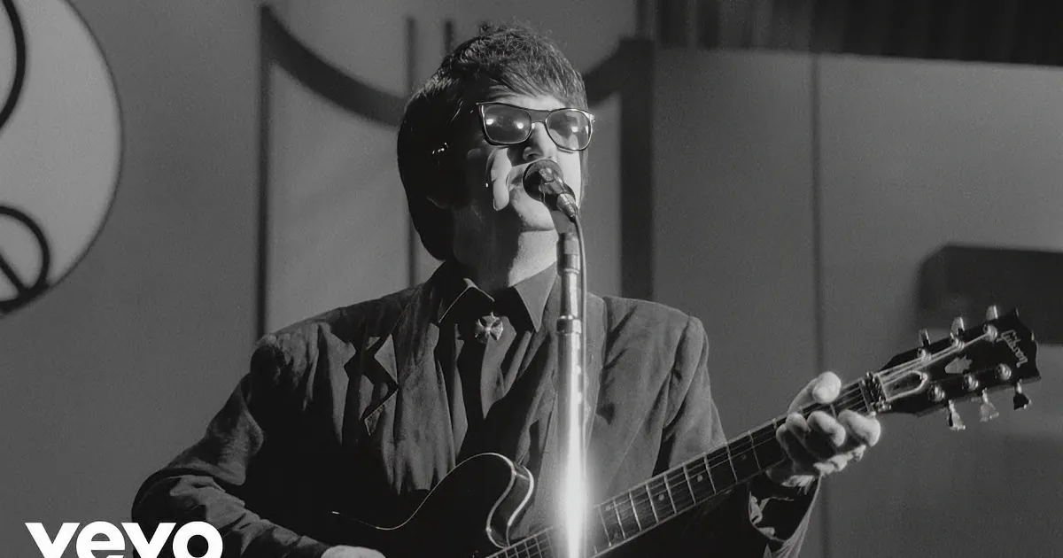 Two musicians play guitars on stage, one in a suit, the other in casual attire, surrounded by microphones and musical...