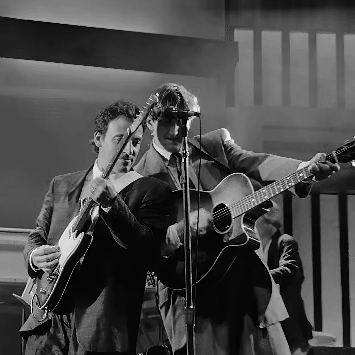 Two musicians in suits perform on stage, one playing guitar while the other sings into a microphone.