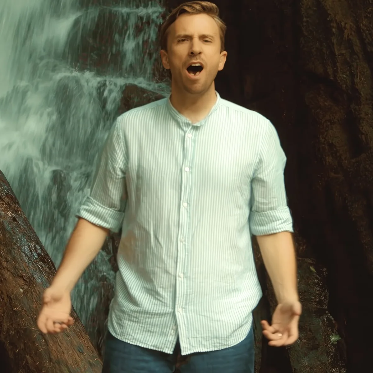 A man in a light blue striped shirt stands with arms outstretched in front of a waterfall.