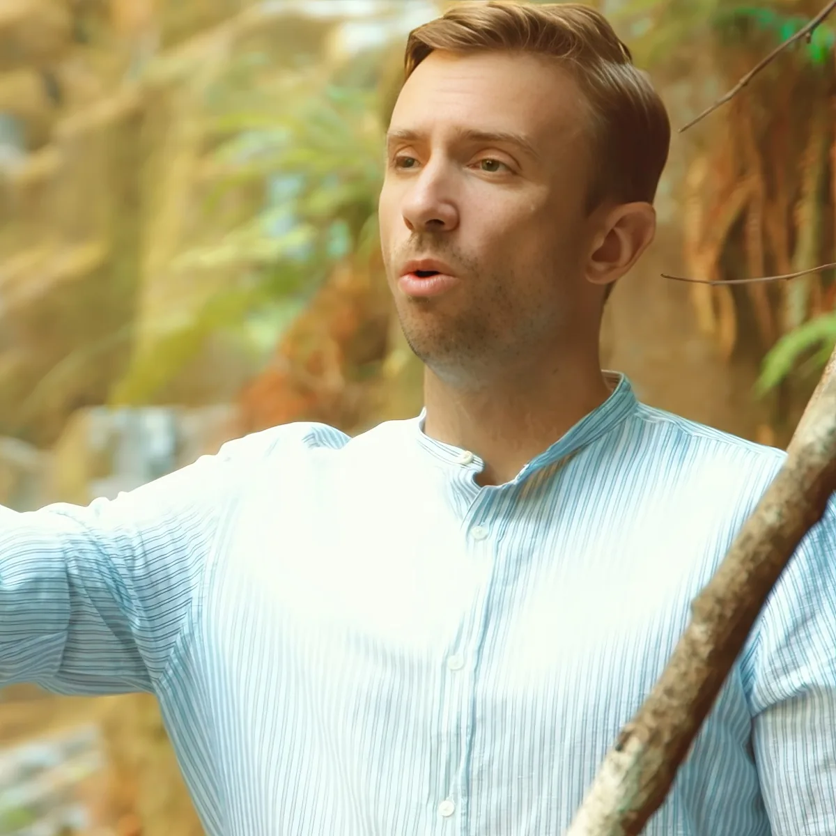 A man in a light blue striped shirt stands outdoors, holding a branch, with a blurred forest background.