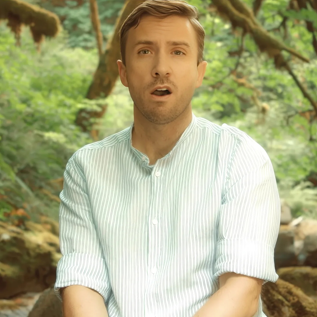 A man in a striped shirt sits outdoors with a forest background, appearing surprised or shocked.