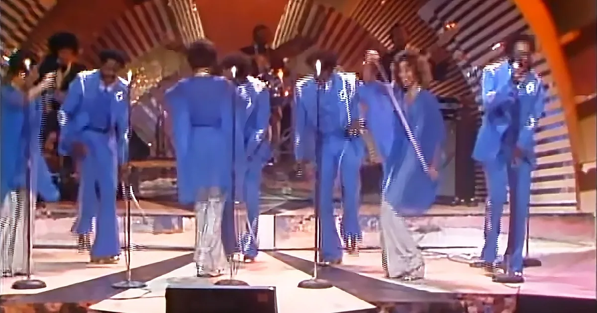 A group of performers in blue suits and silver boots are on stage, singing into microphones.