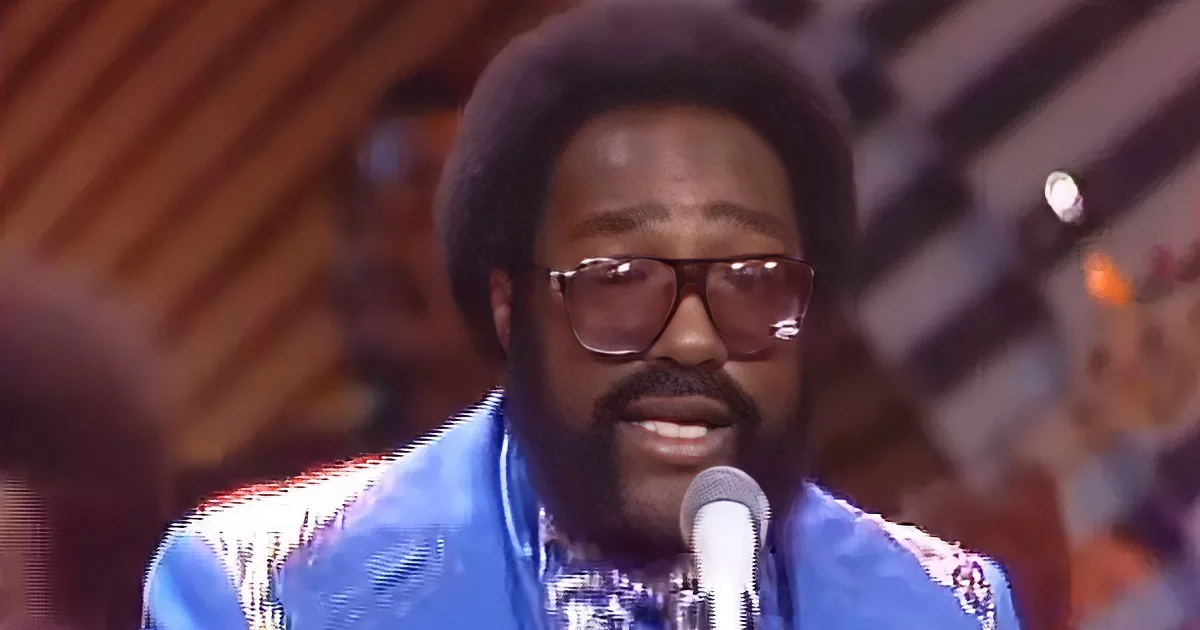 A man with a large afro, glasses, and a blue outfit sings into a microphone.