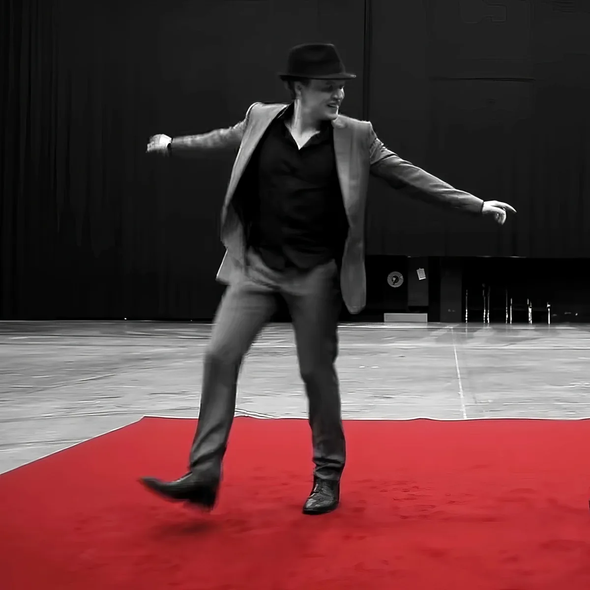 A man in a suit and hat dances on a red carpet in a black-and-white setting.