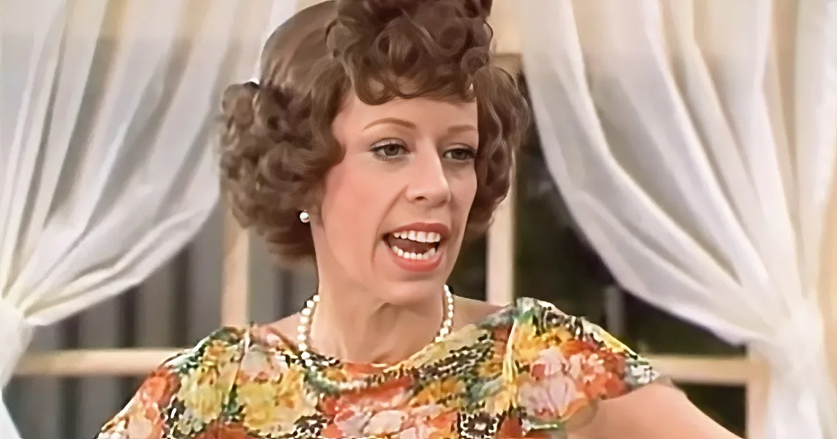 A woman with curly hair, wearing a floral dress and pearl necklace, is speaking with her mouth open.