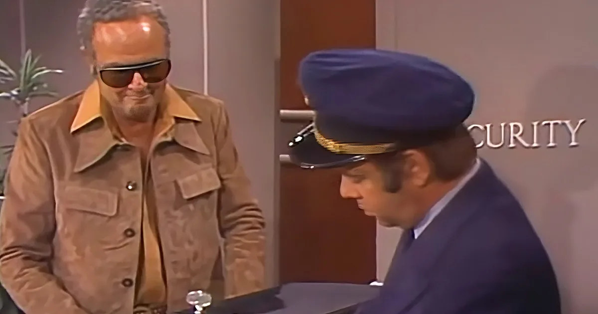 A man in a brown jacket and sunglasses stands beside a security officer in a blue uniform.