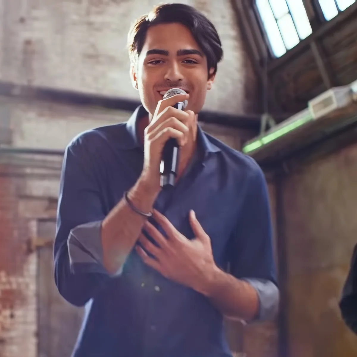 A man in a blue shirt holds a microphone, singing with his hand on his chest in a rustic setting.