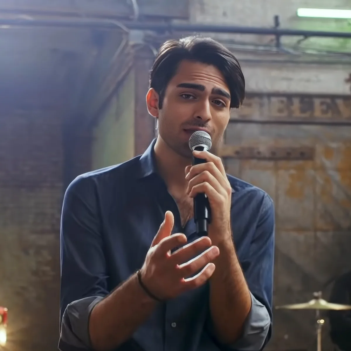 A man in a dark shirt holds a microphone, gesturing with his other hand, in a dimly lit setting.