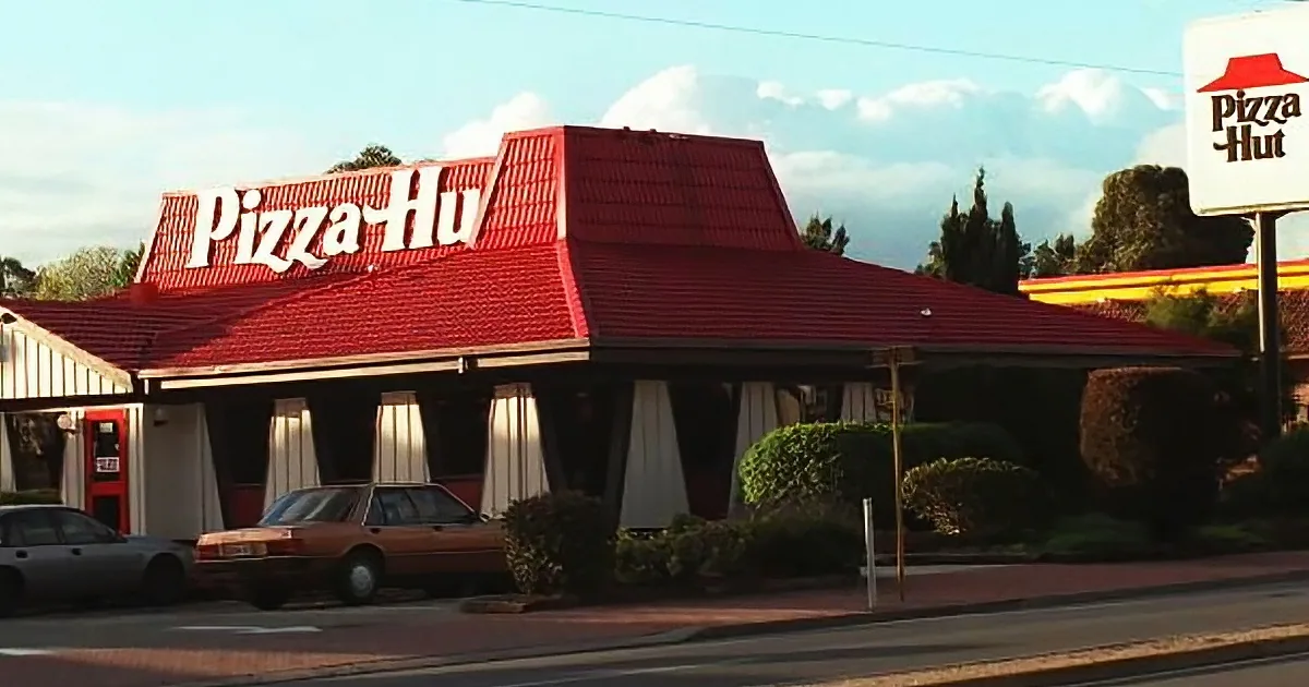 Pizza Hut restaurant with red roof and sign, cars parked, bushes, and clear sky.