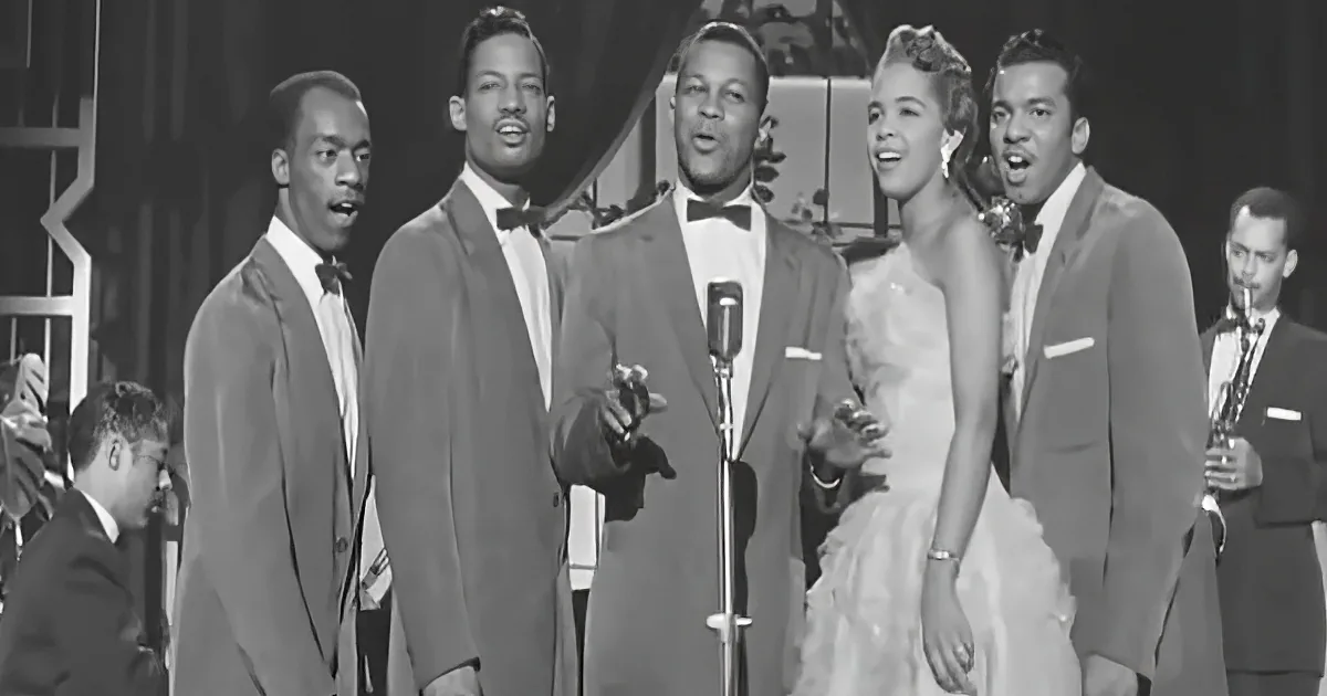 A group of five performers in formal attire sing on stage, accompanied by a saxophonist.