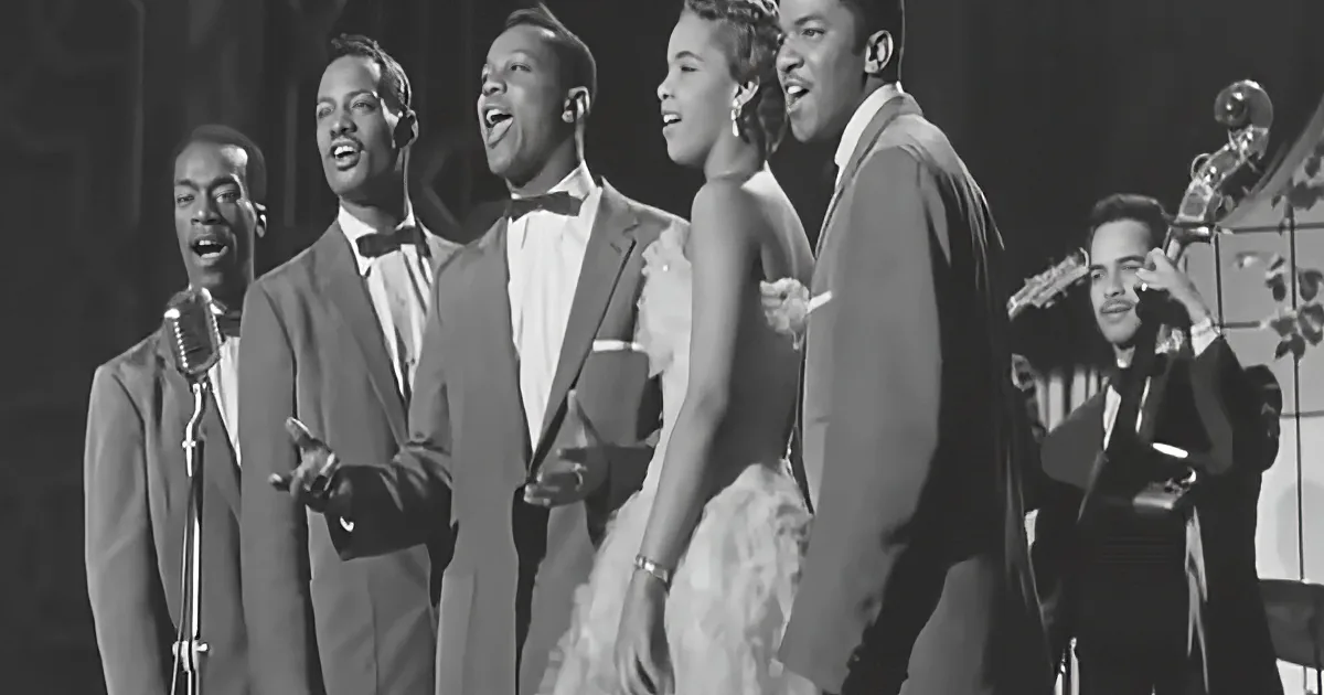A group of five performers in formal attire sing on stage, accompanied by a musician playing a bass.