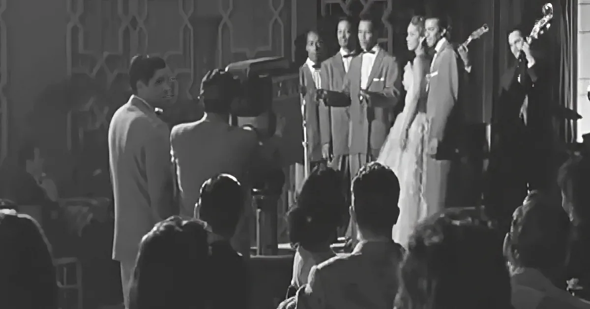 A group of men in suits performs on stage, with a woman in a dress and a musician playing a trumpet.
