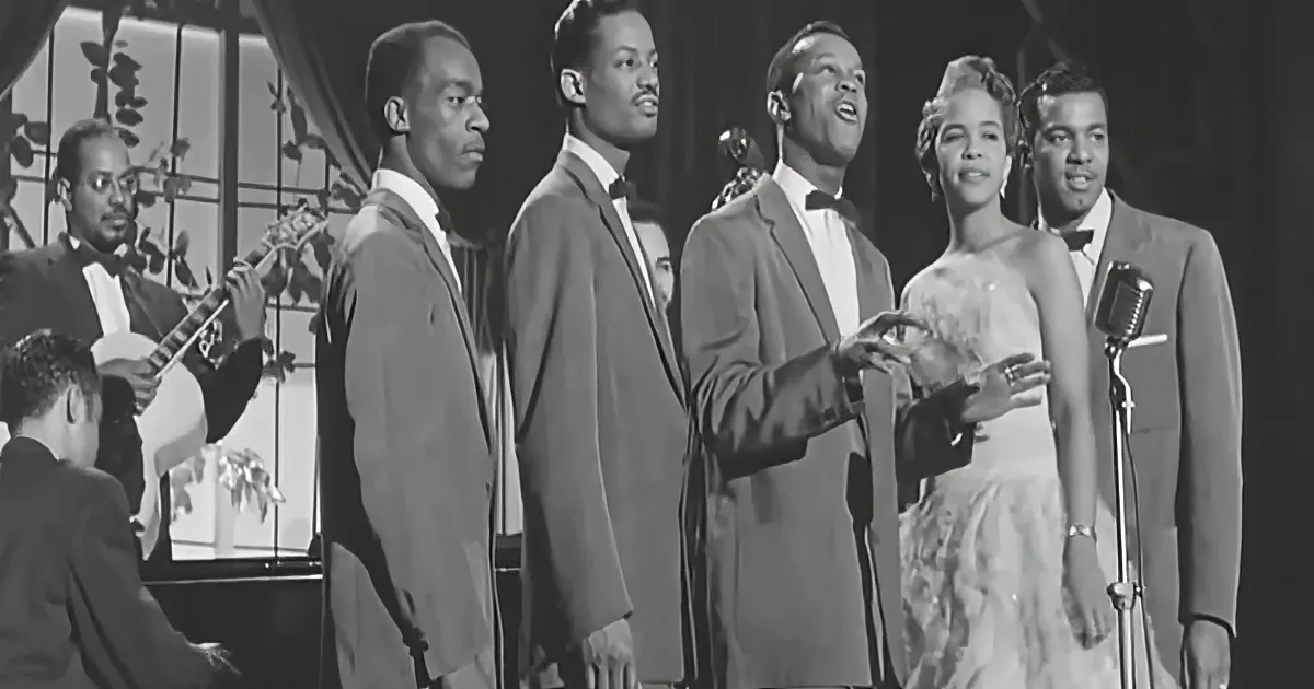 A group of six people in formal attire singing on stage, accompanied by a musician playing a string instrument.