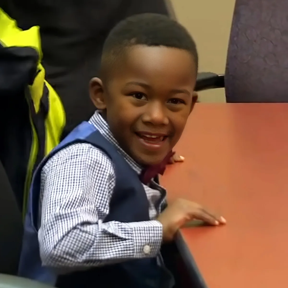 A young boy in a blue vest and checkered shirt smiles while leaning on a red surface.
