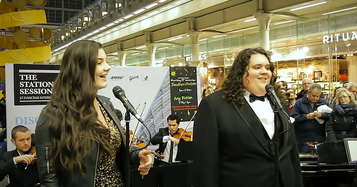 A woman in a black leather jacket sings into a microphone while a man in a tuxedo smiles beside her.
