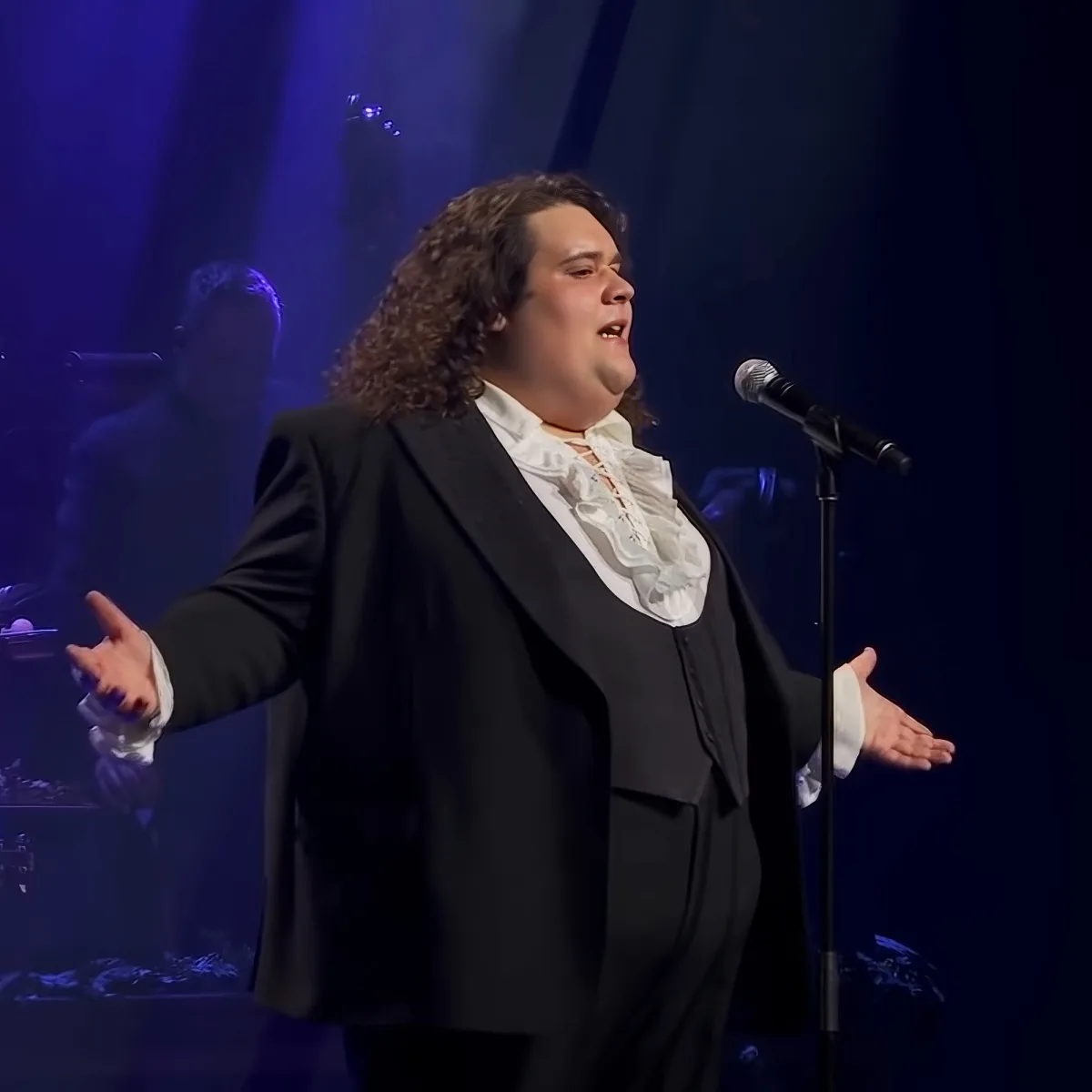 A person in a black suit with a white ruffled shirt sings on stage with a microphone, arms outstretched.
