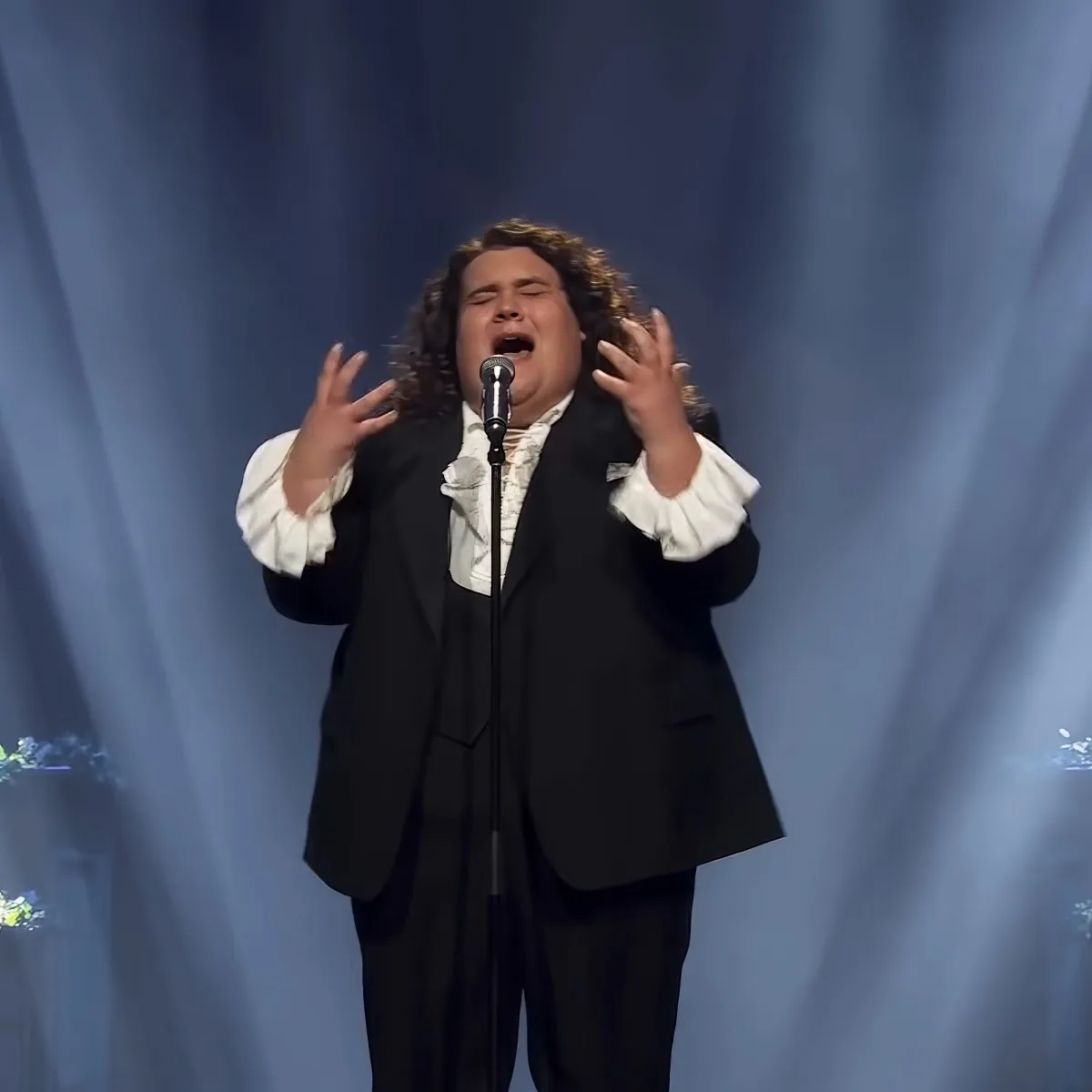 A person in a black suit and white shirt sings passionately on stage with arms raised.